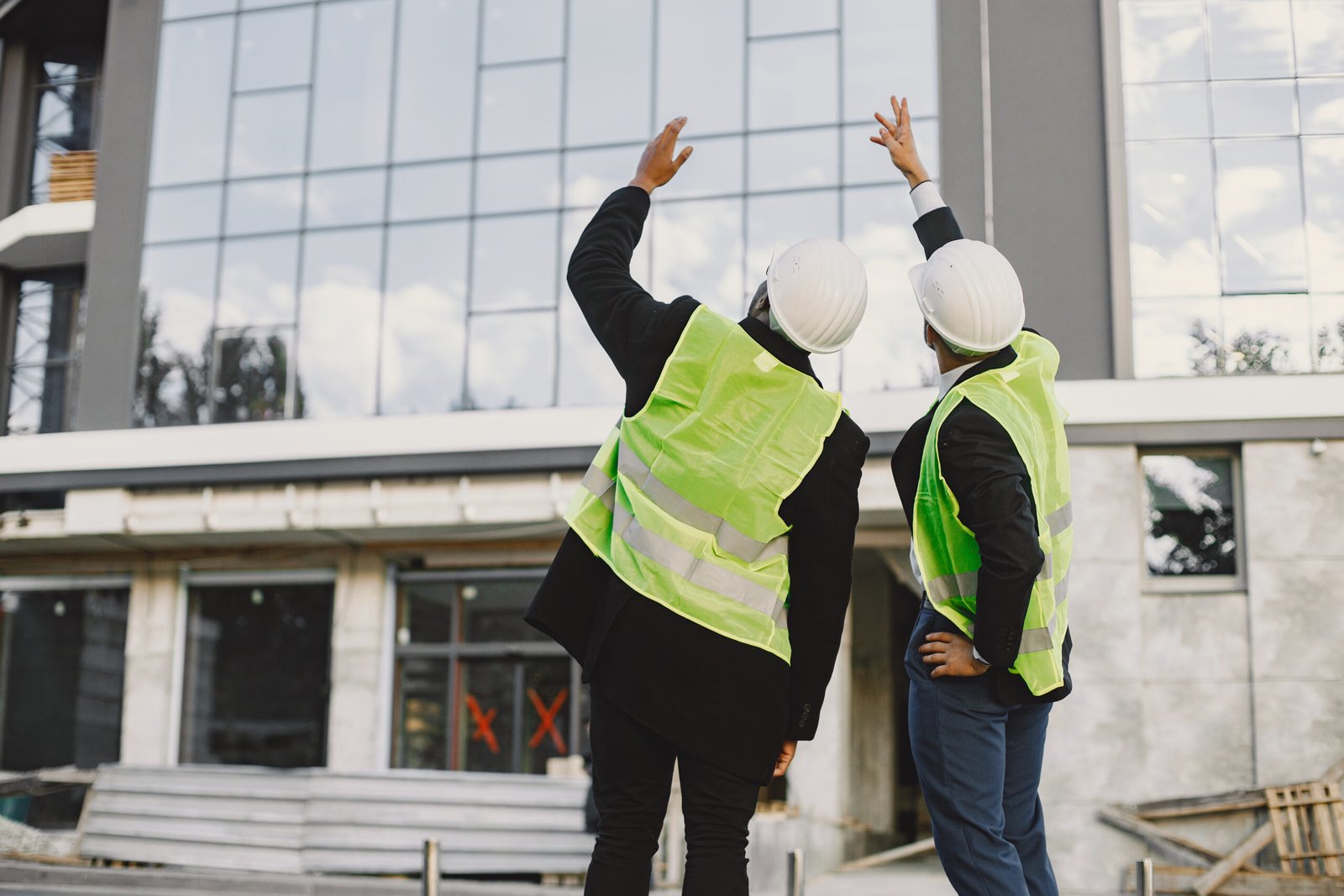 Multi racial builders standing outdoors, back view. Wearing uniform, talking about new glass building. Working on the poject. City infrastructure. Black man.